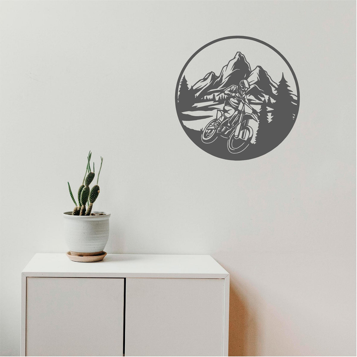 Dark grey circular mountain biker wall decal with mountains and pine trees above a white cabinet beside a cactus.