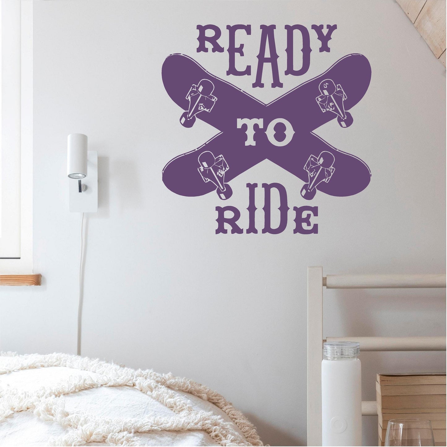 Purple Ready to Ride wall decal with two crossed skateboards above a bed on a white wall.