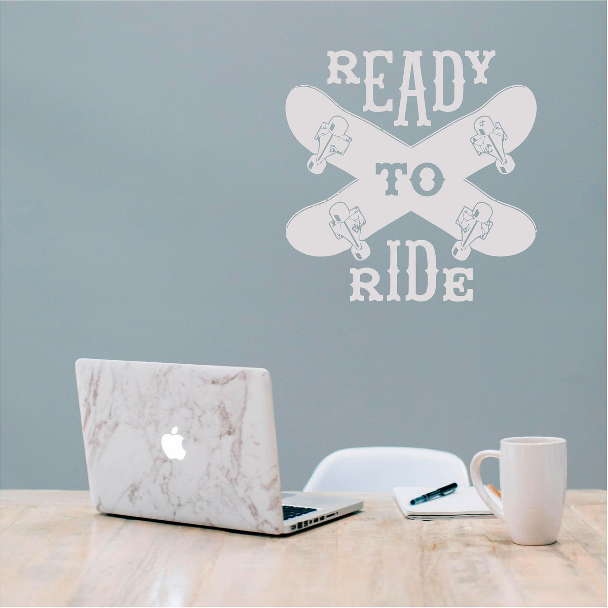 White Ready to Ride wall decal with two crossed skateboards on a muted blue wall above a desk.