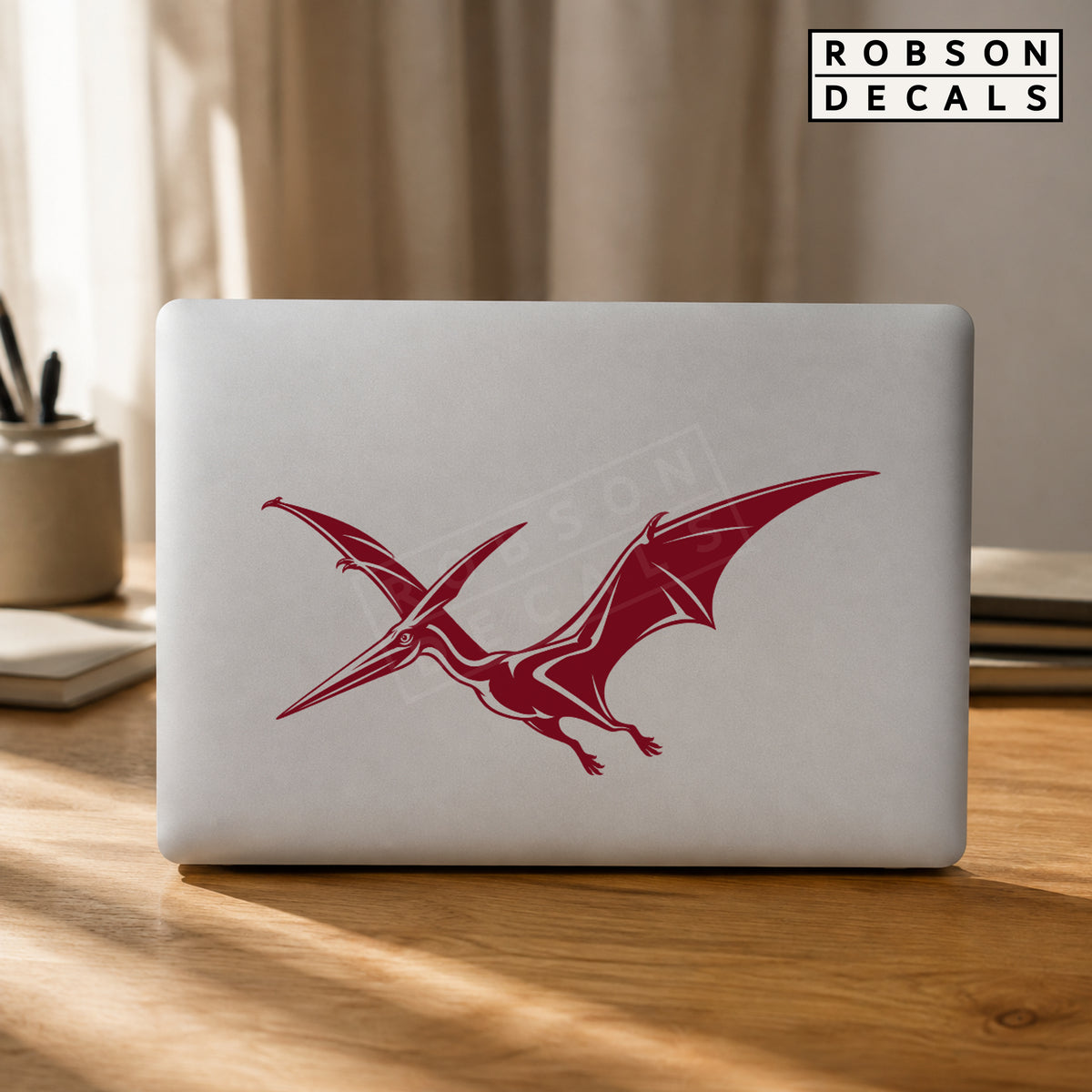Burgundy flying pterodactyl decal on a silver laptop lid, styled Robson Decals mockup